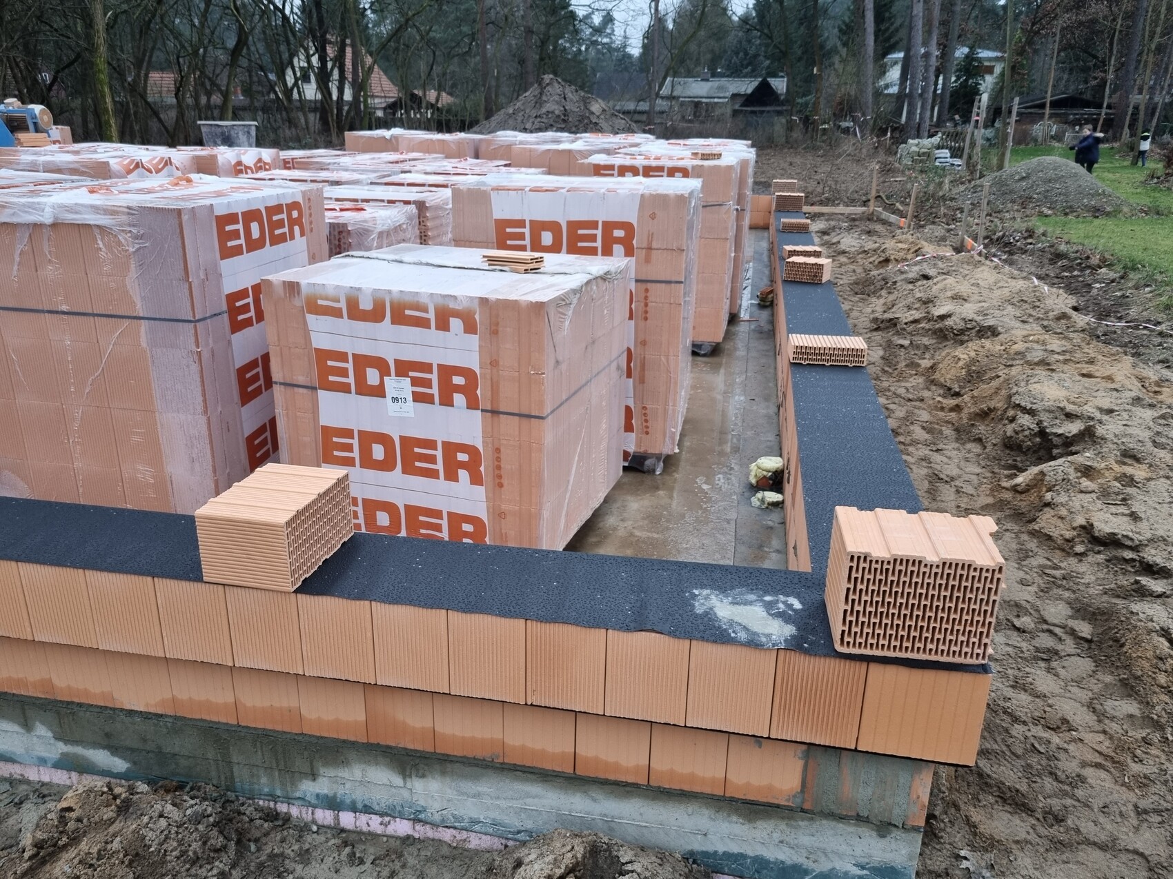 Baustelle mit vielen EDER-Ziegelverpackungen, orange Lochziegel-Wand am Fundament und schwarzer Dichtbahn.