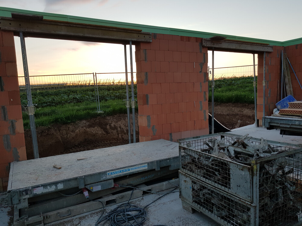 Rohbau aus roten Ziegeln mit offenen Türen, Bauwerkzeuge und Korb voller Metallteile; Abendlicht.