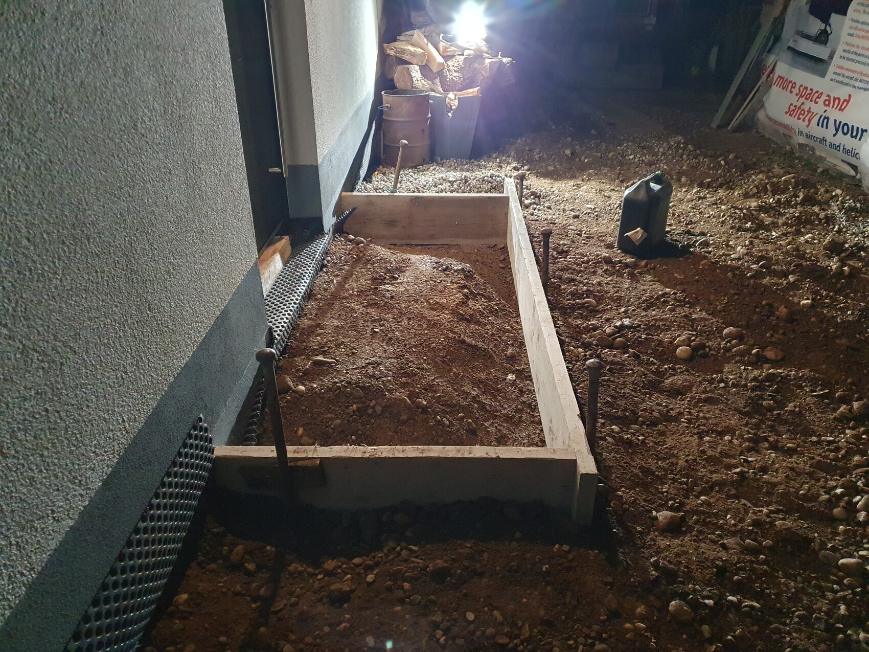 Fundament-Schalung aus Holz im Erdgraben neben der Hauswand; Erde, Kies und Werkzeuge sichtbar.
