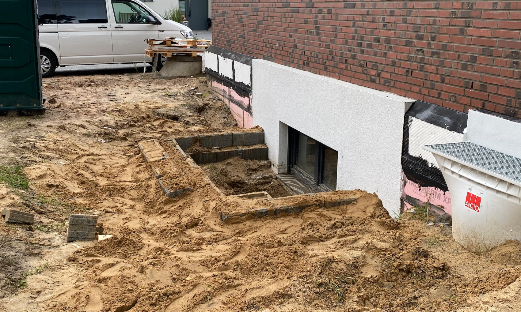 Baustelle am Haus: offenes Kellerfenster, Erdarbeiten, Sandhügel, Paletten links.