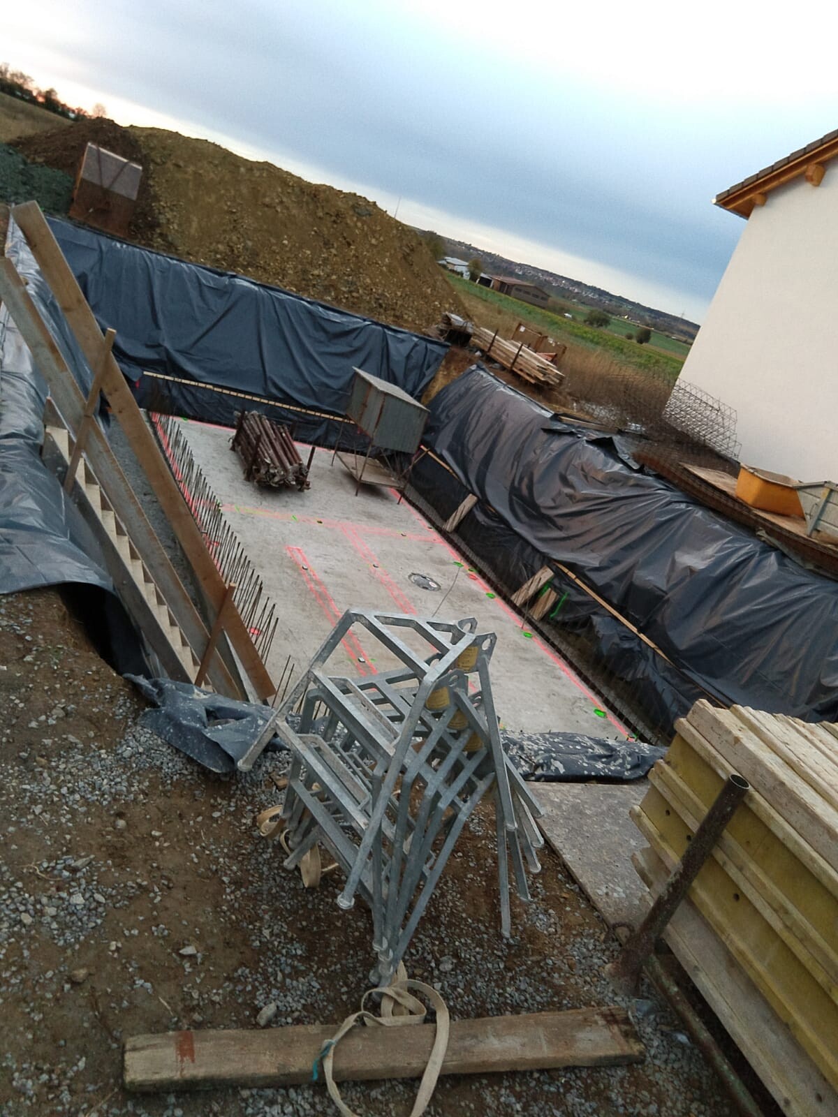 Baustelle mit Baugrube, Betonplatte, Stahlrahmen, Holzgerüsten und schwarzer Baufolie.
