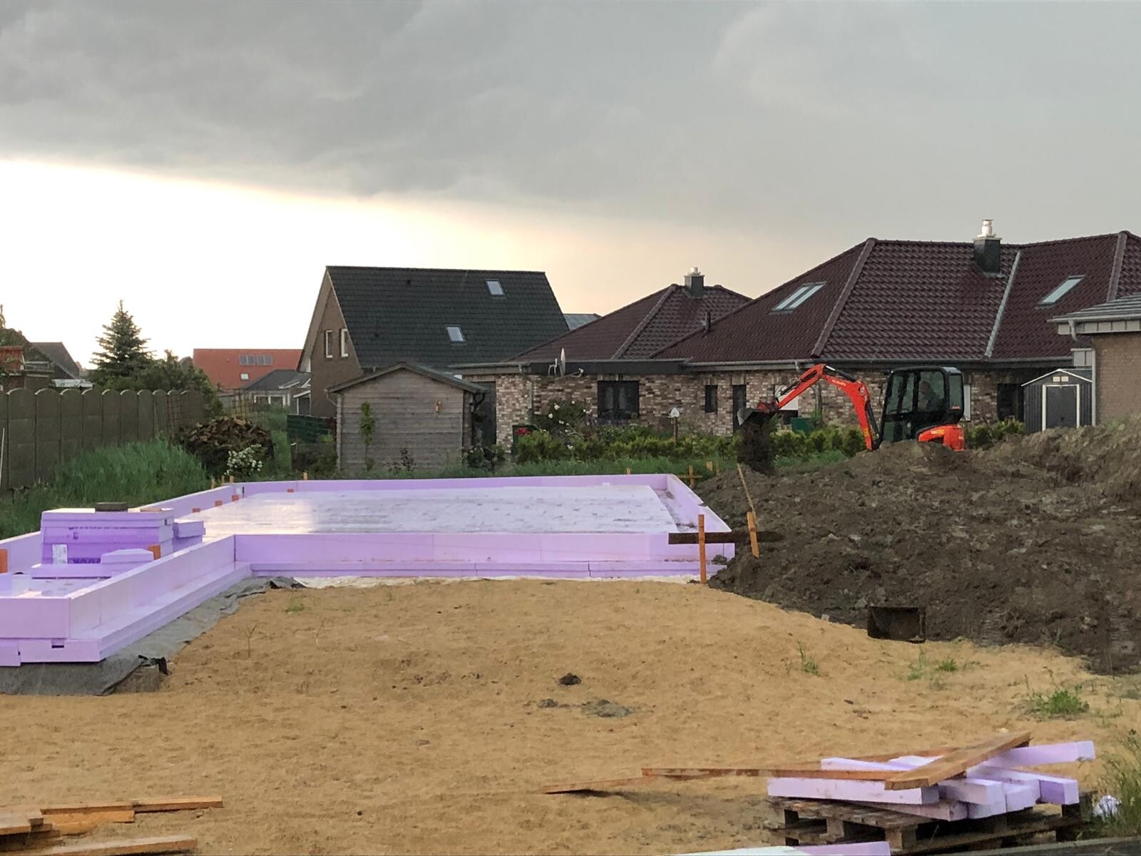 Baustelle mit lilafarbenen Fundament-Isolierplatten, Bagger im Hintergrund und Wohnhäusern.