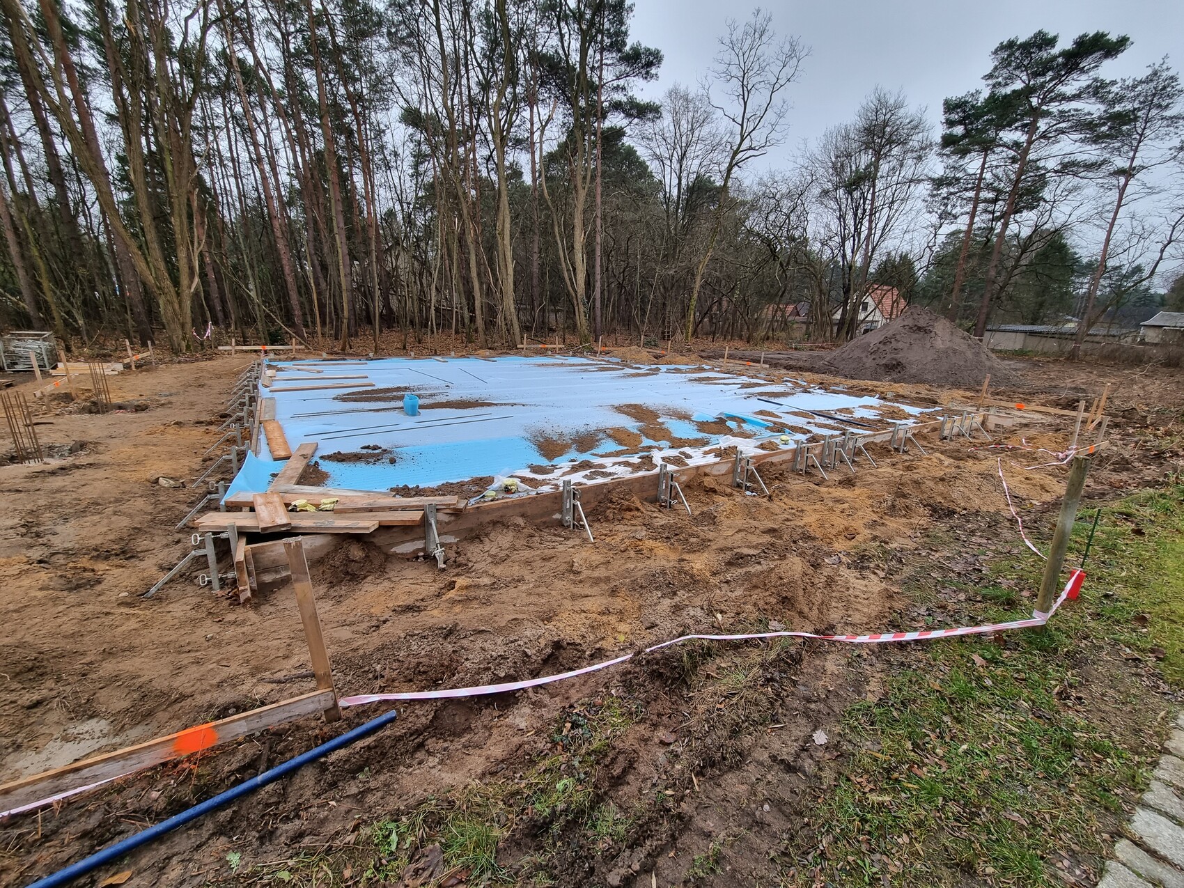 Fundamentplatte wird vorbereitet: blaue Abdeckung, Holzformen, Erde, Bauabsperrband, Wald.