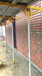 Baustelle mit Gelb-Lila Gerüst neben roter Backsteinfassade, Erdreich sichtbar