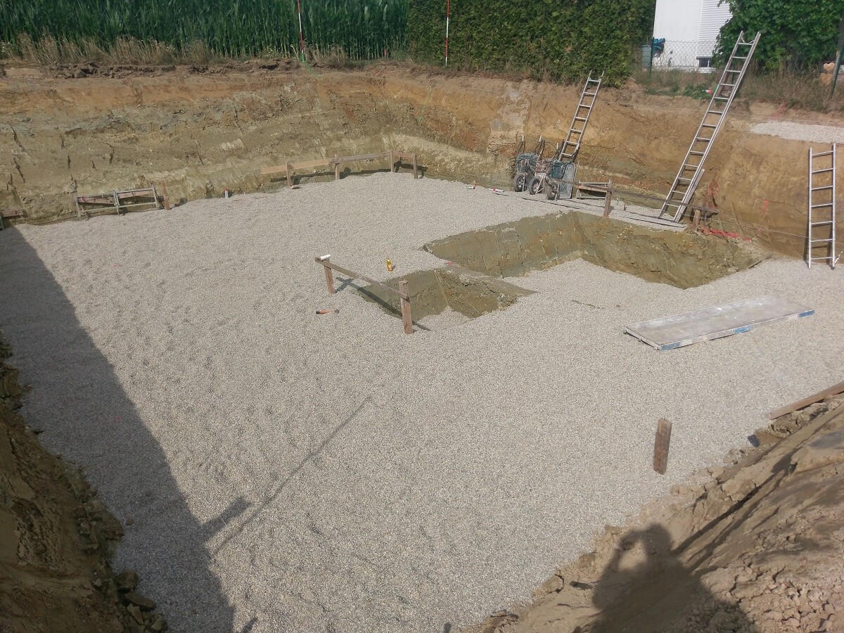 Baugrube mit Kiesboden, mehreren Leitern, Holzstützen und Baumaschinen auf einer Baustelle.