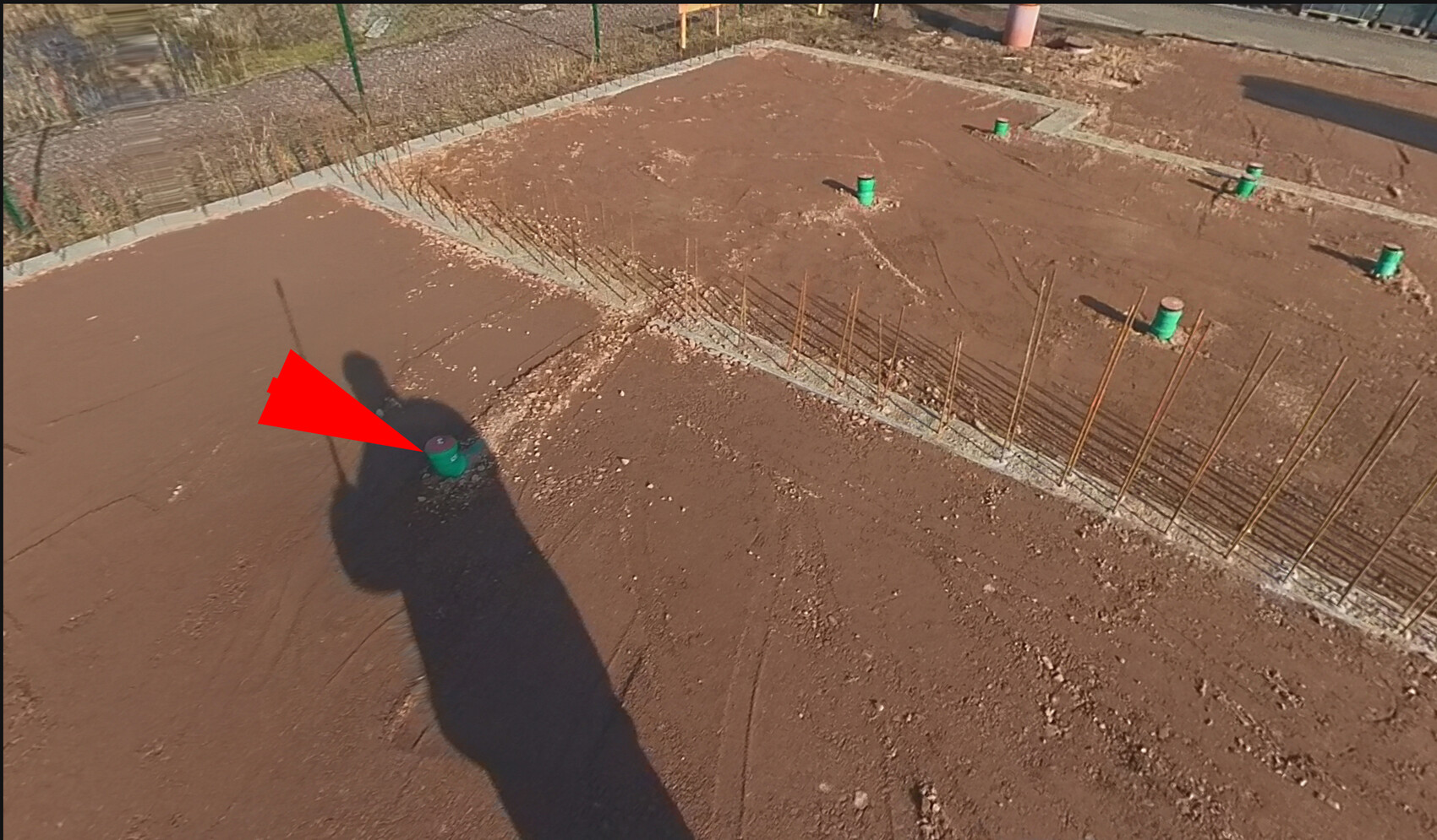 Baustelle mit Erdreich, sichtbare Bewehrungsstangen, grüne Rohrstücke, Schatten einer Person.