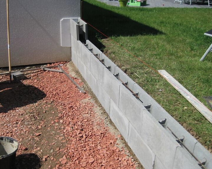 Baustelle mit halbfertiger Betonsteinmauer und Bewehrung am Fundamentrand.