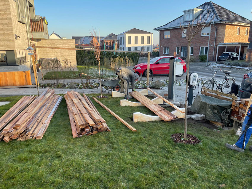 Person bearbeitet Holz im Vorgarten; Holzbretterstapel, Bauholz, Wohnhäuser im Hintergrund.