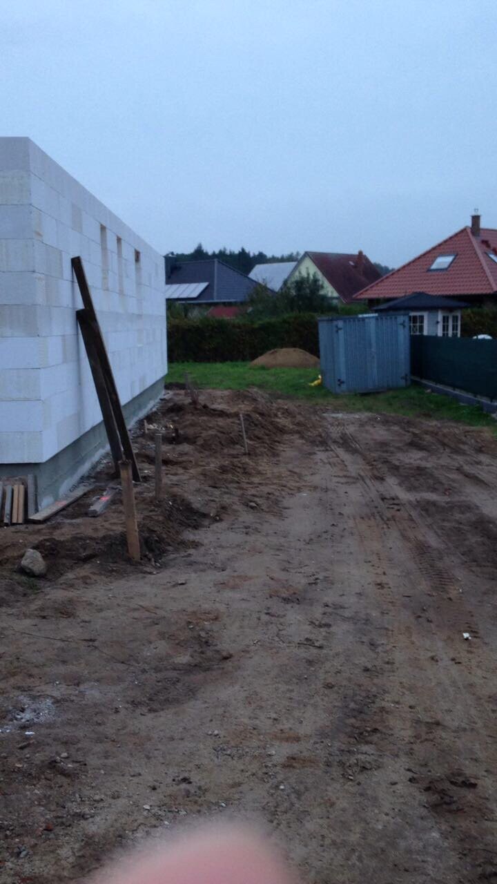 Baustelle: unfertiges Haus aus weißen Blöcken, Holzpfosten; blauer Container, Häuser dahinter.