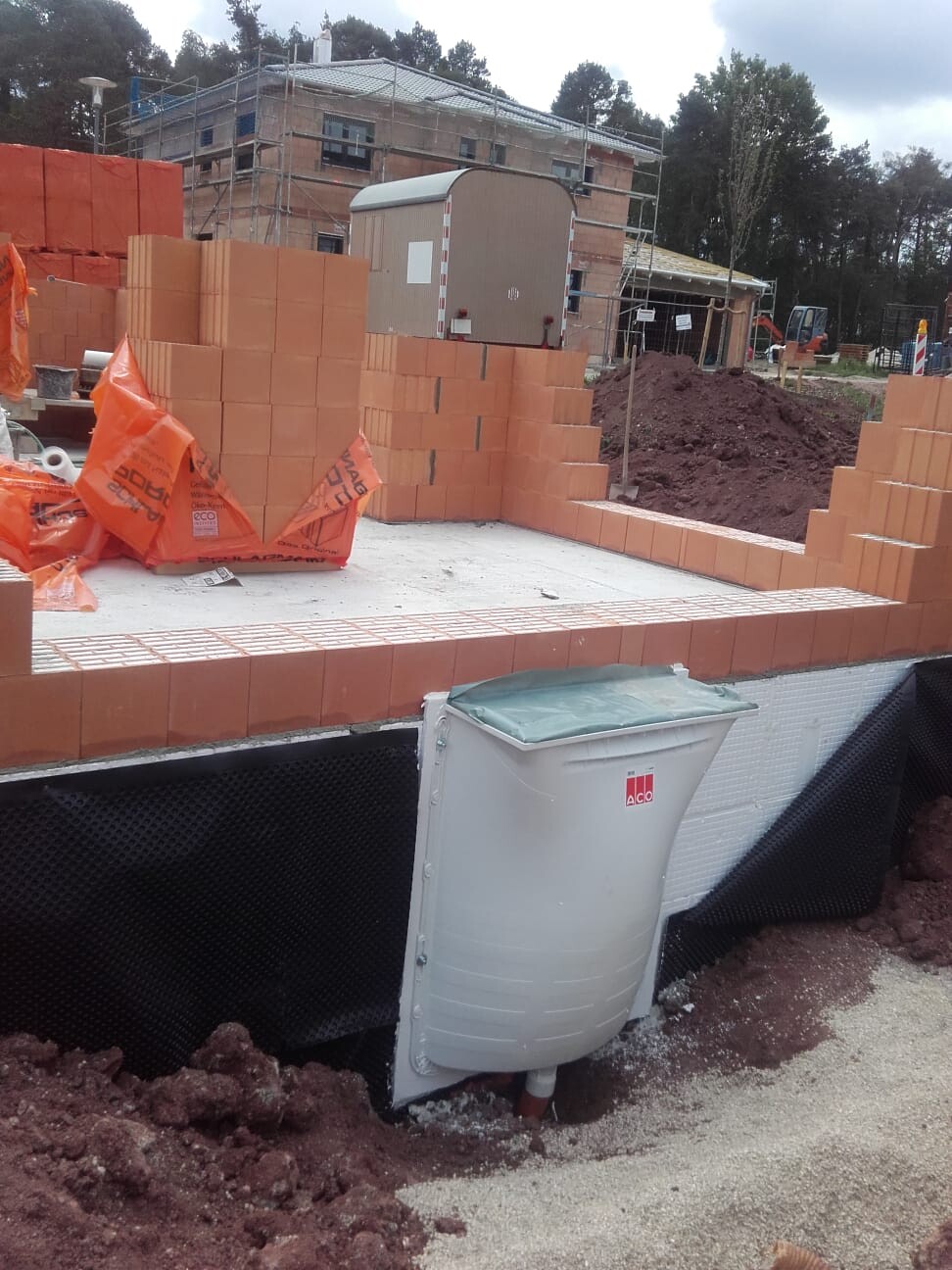 Baustelle mit gestapelten Ziegeln, Baucontainer, Gerüste und Erdaushub im Vordergrund.