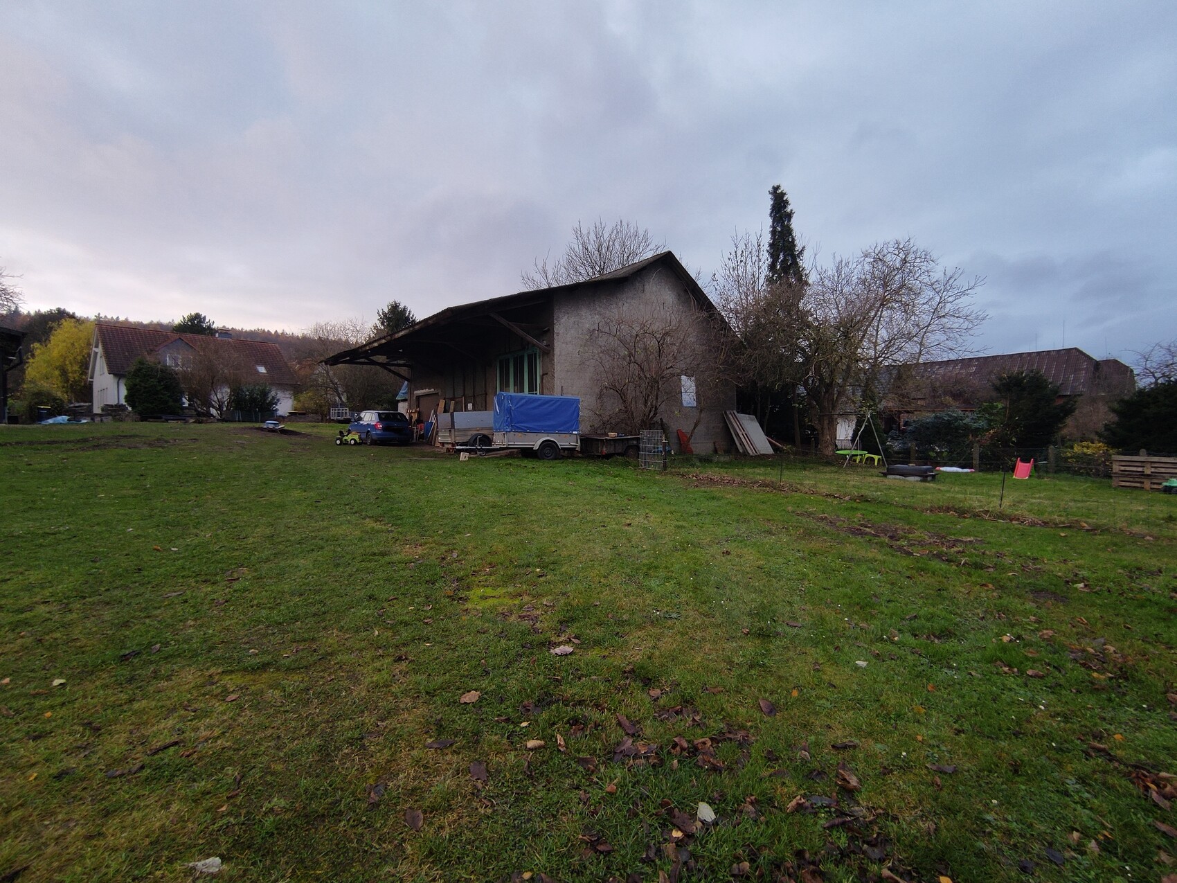 Rohbau-Halle auf gruenem Grundstueck mit Garten, Fahrzeugen und Bauutensilien.