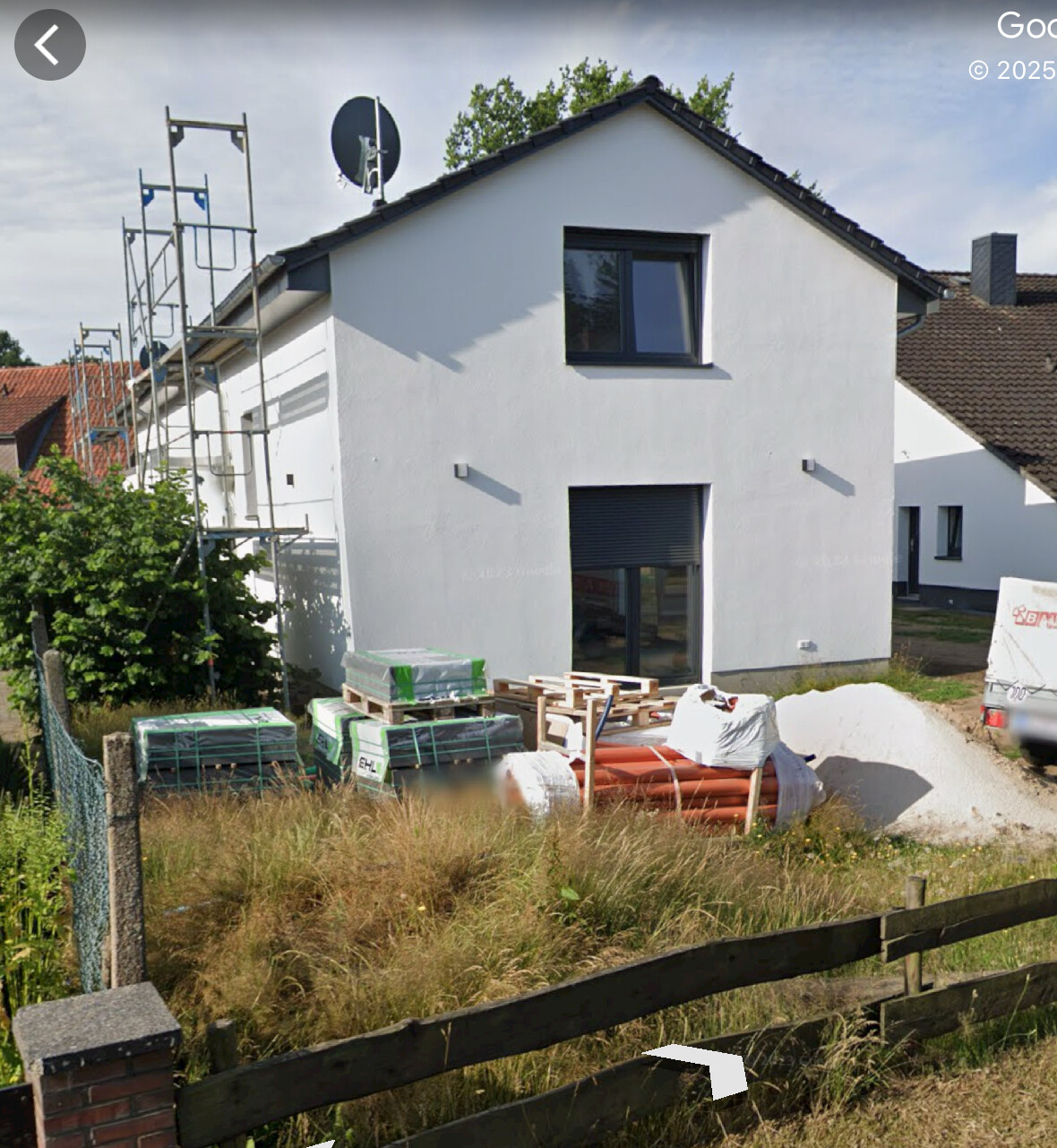 Weißes Einfamilienhaus im Bau mit Gerüst links; Baustellenmaterialien im Vorgarten.