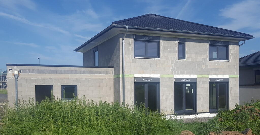 Zweistöckiges Neubauhaus aus Betonblöcken mit großen Fenstern und grünem Umfeld.