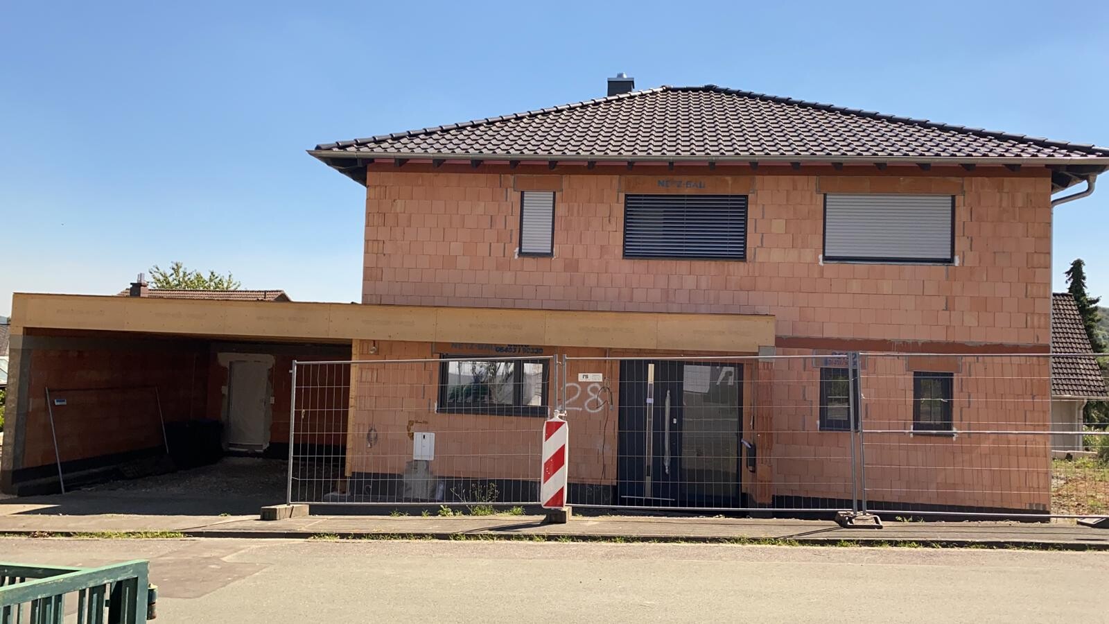 Zweistöckiges Haus im Rohbau mit Backsteinfassade, Bauzaun davor und Carport links.