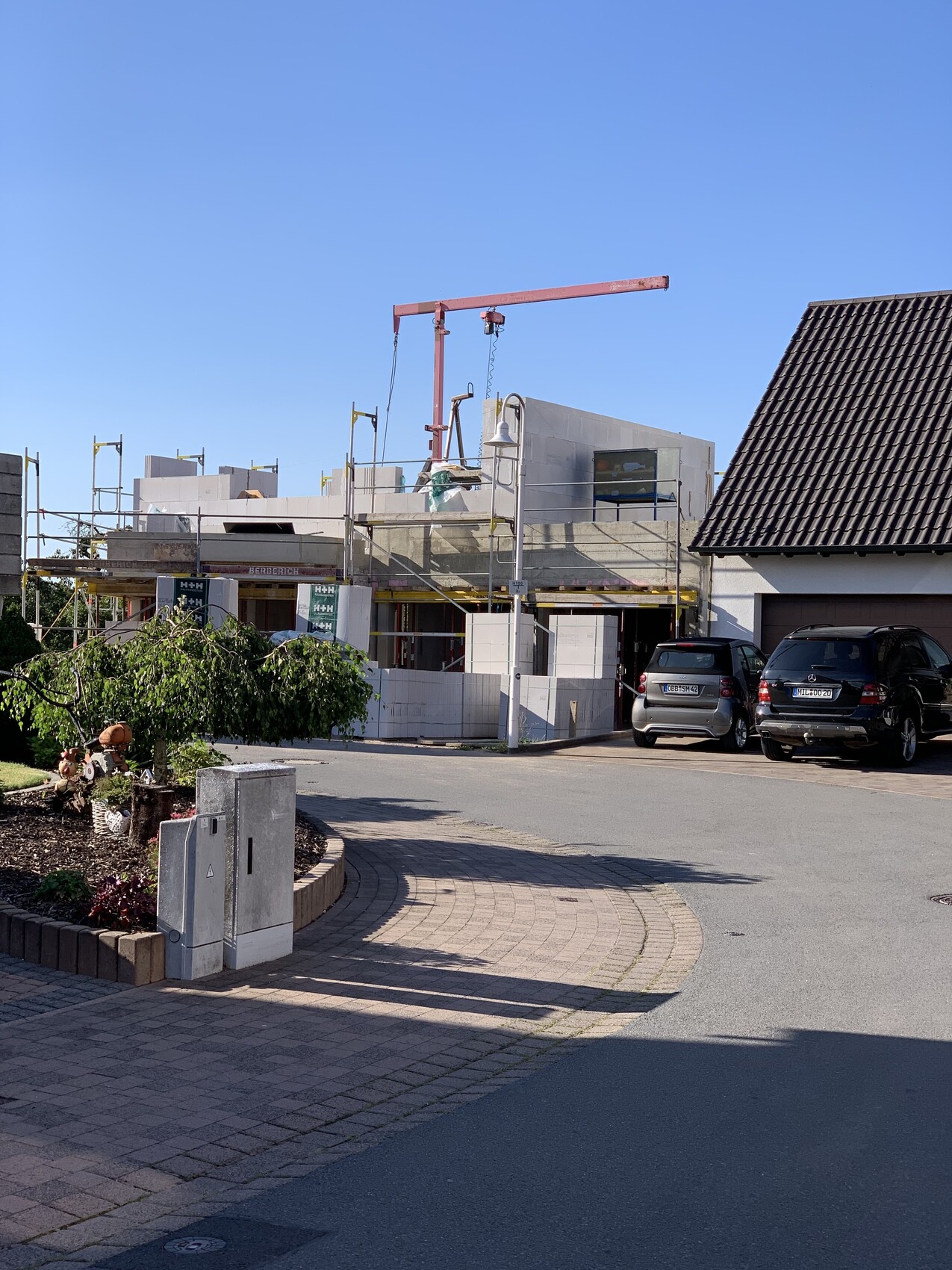 Rohbau eines Hauses mit Gerüst, rotem Kran; daneben zwei geparkte Autos.