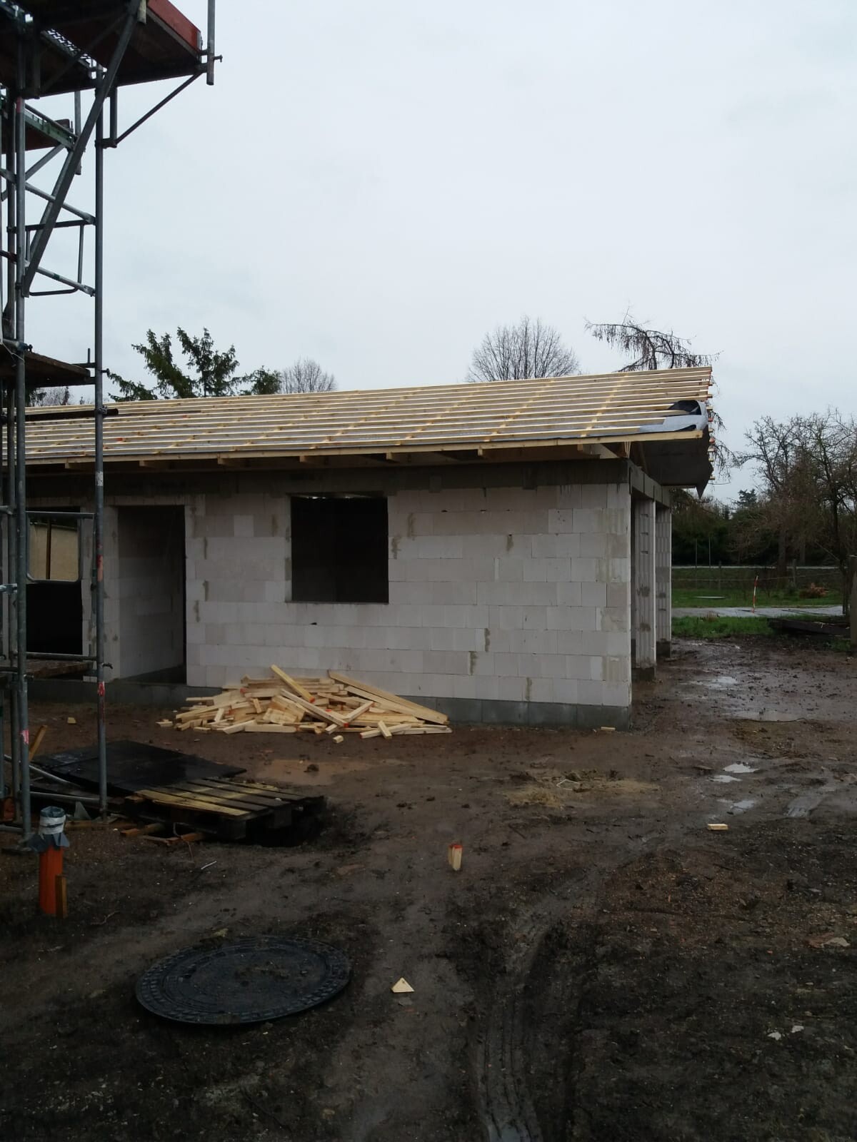 Kleines Gebäude aus Betonblöcken, Dachkonstruktion aus Holz, Holzreste am Boden, links Gerüst.
