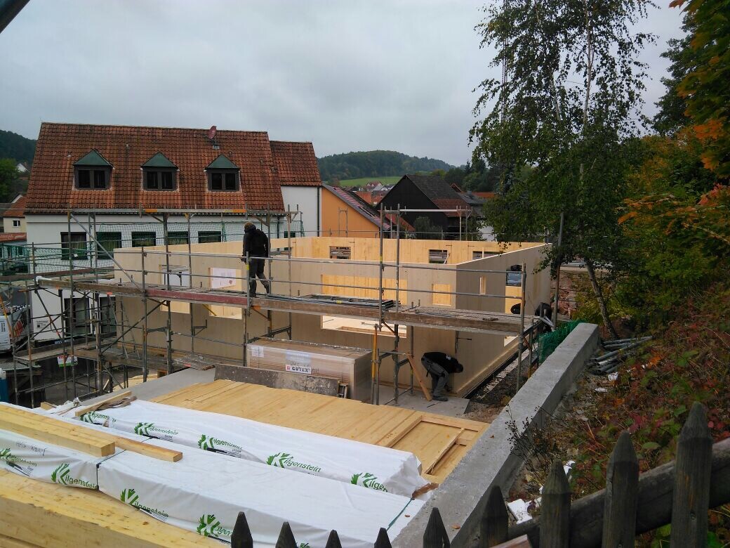 Baustelle mit Holzwänden, Gerüst und Arbeiter; umliegende Häuser und Bäume.