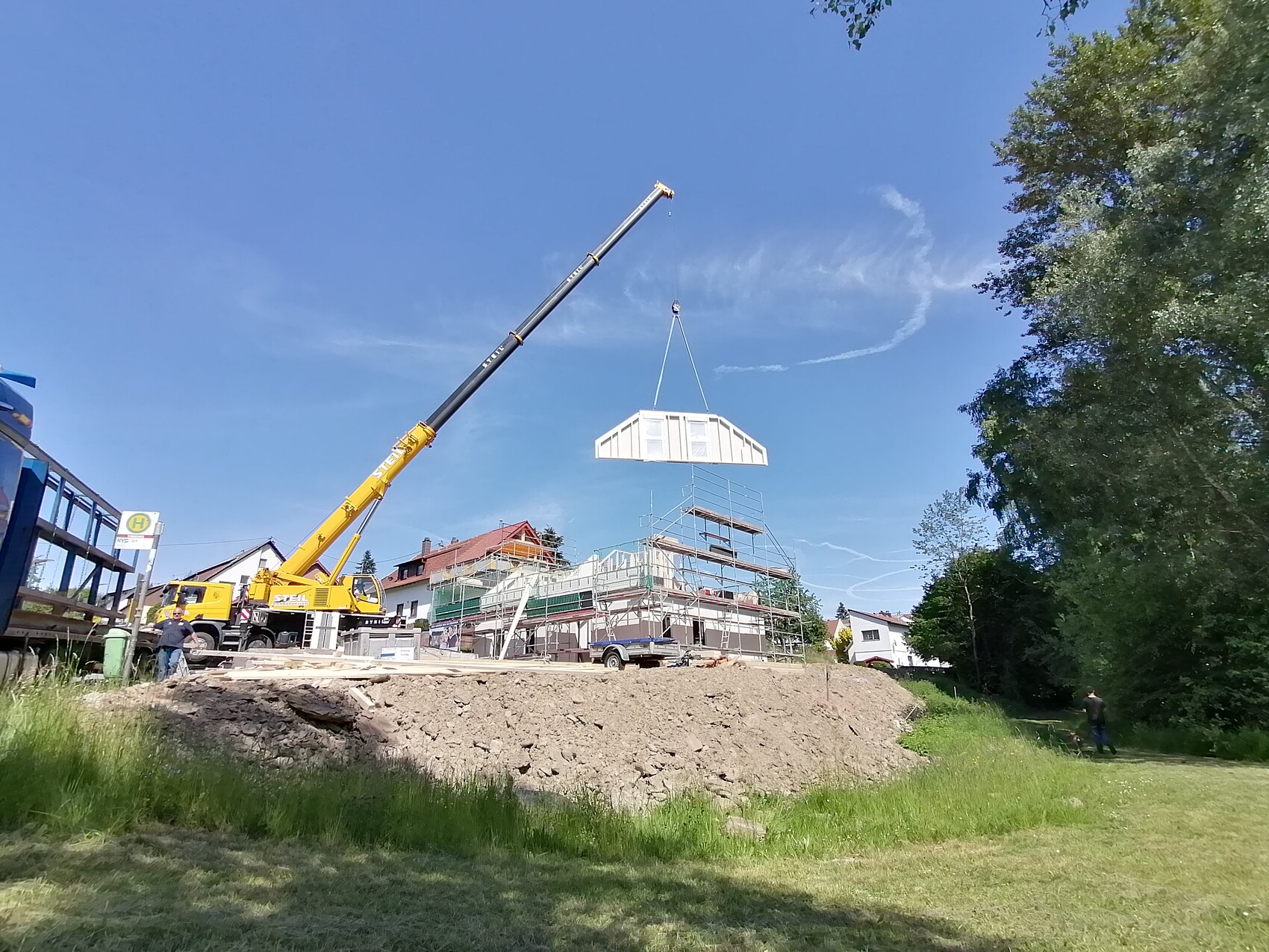 Baukran hebt vor dem Rohbau eine weiße Fertigteilwand über Gerüst; Baustelle mit Gras und Bäumen.
