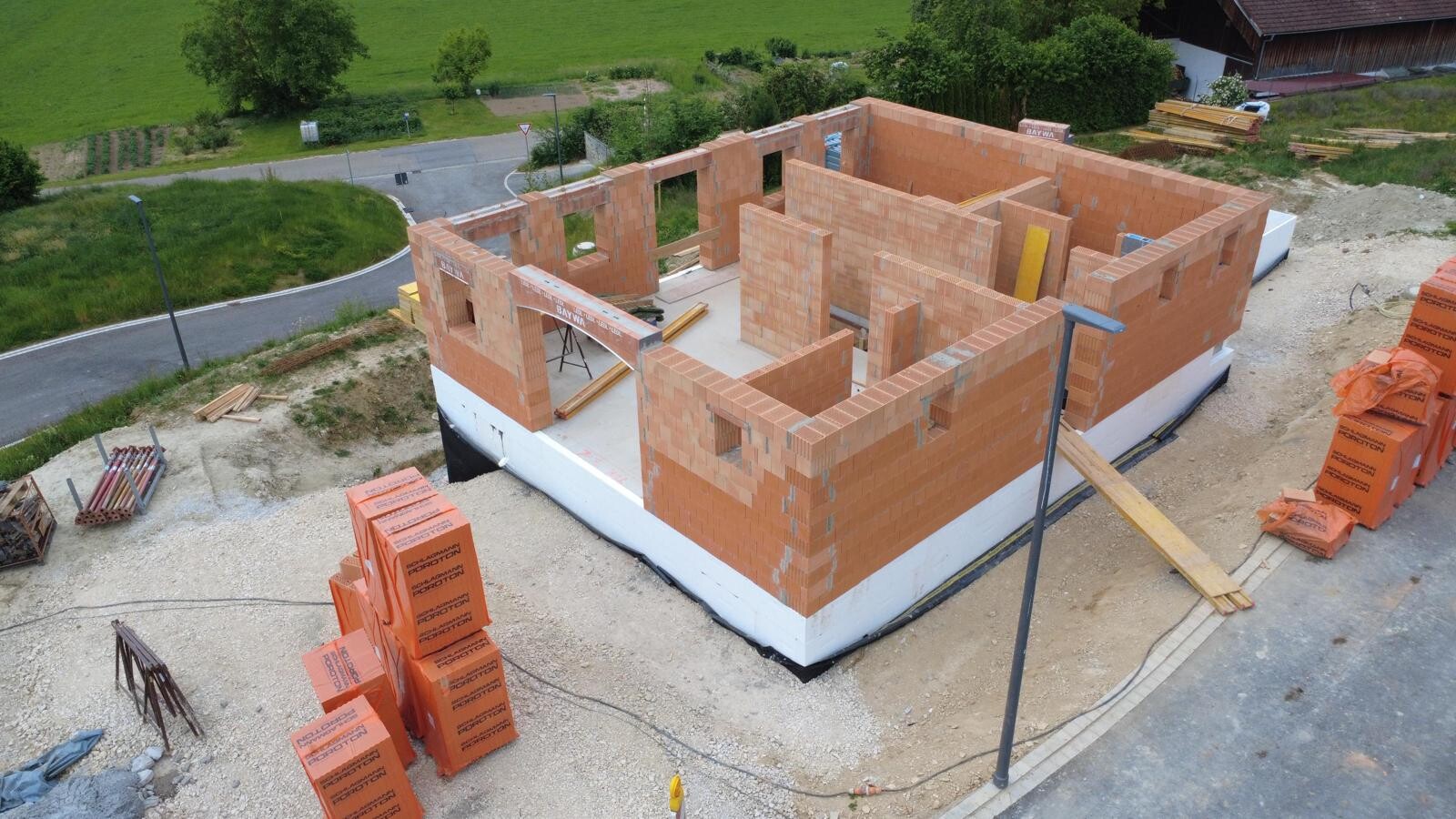 Rohbau aus roten Ziegelsteinen auf Fundament, Innenwände sichtbar, Baustelle mit Holzstapeln.