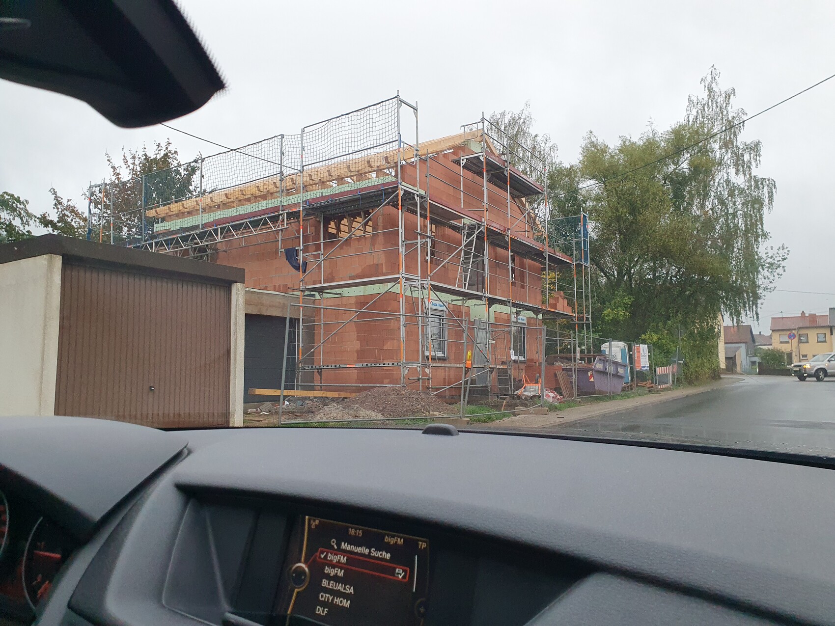 Rotes Ziegelhaus im Bau mit Gerüst; Straßenszene und Bäume, Auto-Dashboard sichtbar.