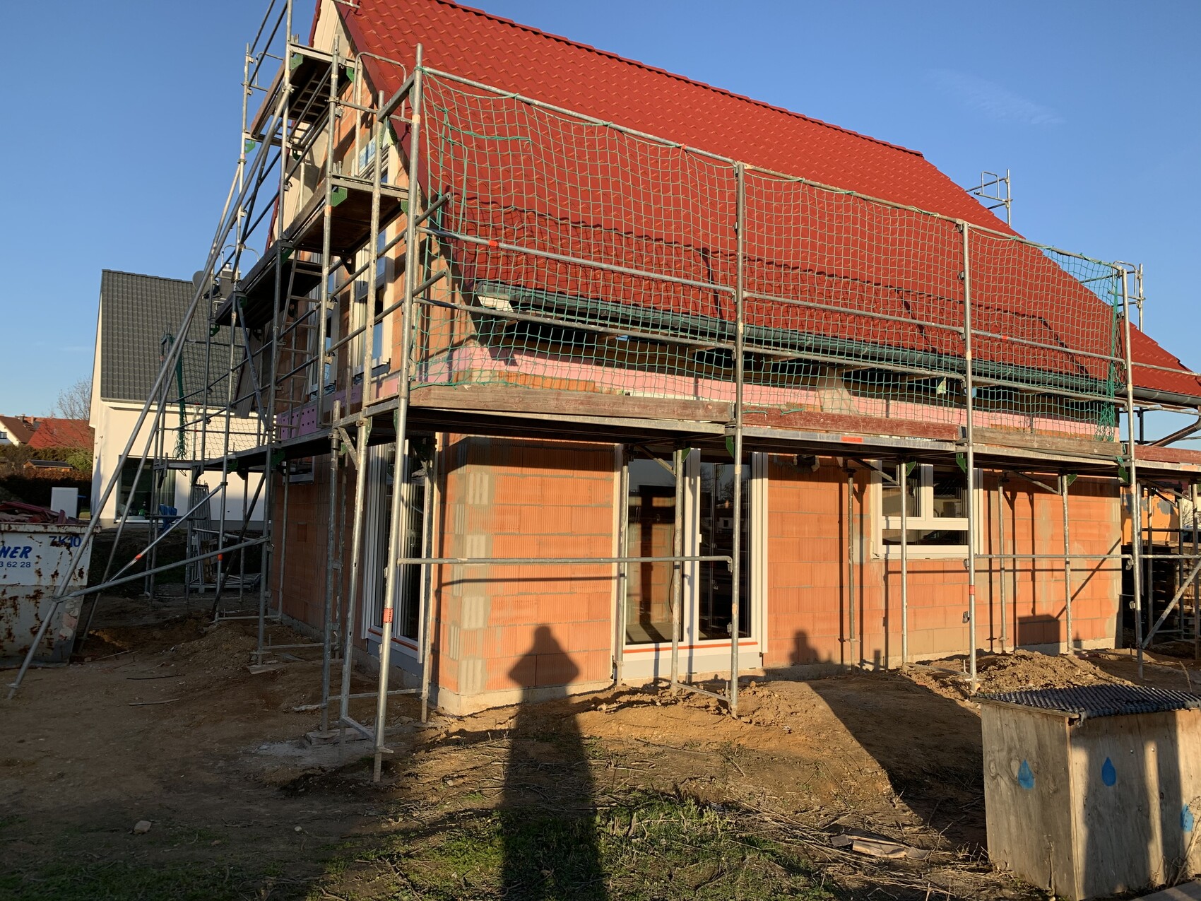 Haus im Bau mit Gerüst, rotem Ziegeldach und orangefarbenen Ziegelwänden; Bauarbeiten sichtbar.