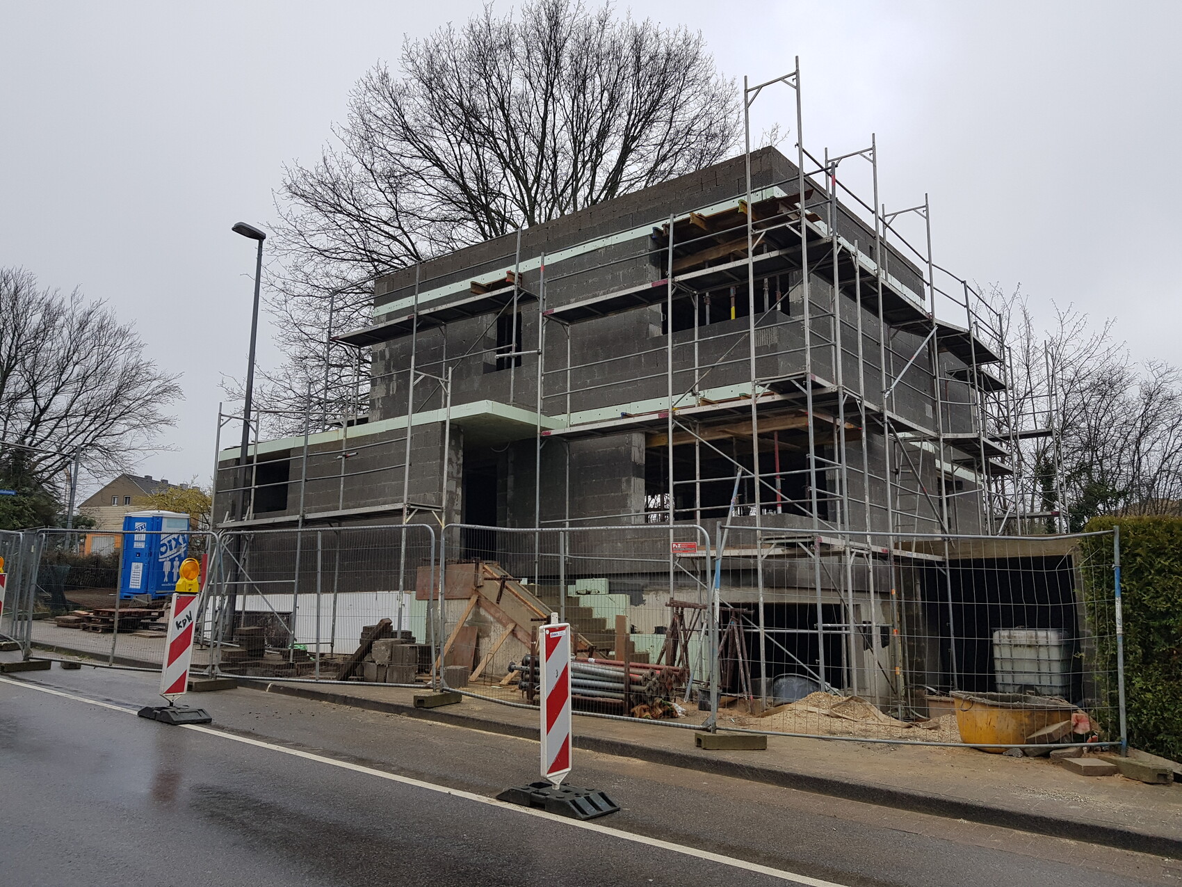 Baustelle: mehrstöckiges Betongebäude mit Gerüst, Absperrungen und Baumaterial am Straßenrand.