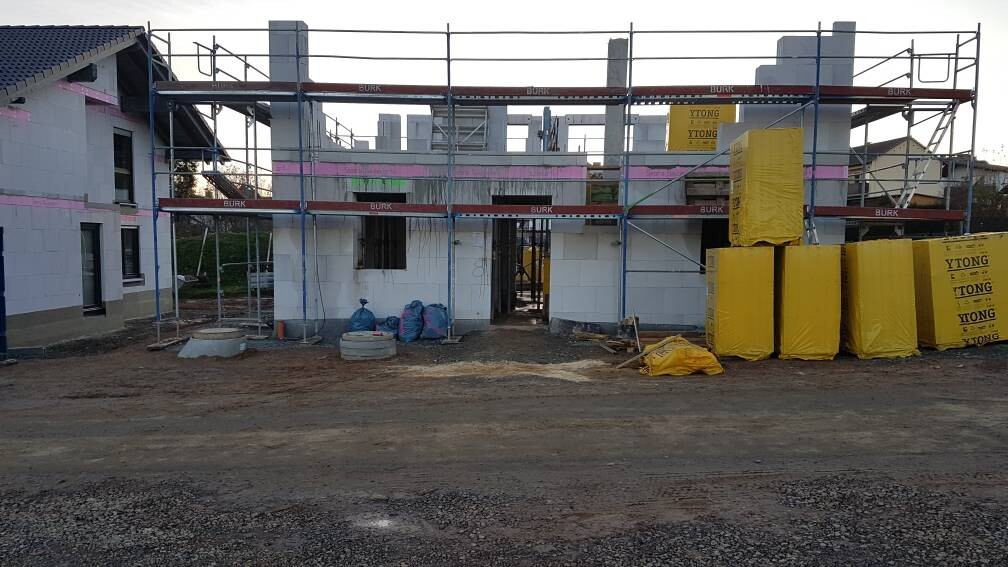 Baustelle: Haus im Rohbau mit Gerüst; rechts gelbes Dämmmaterial gestapelt.