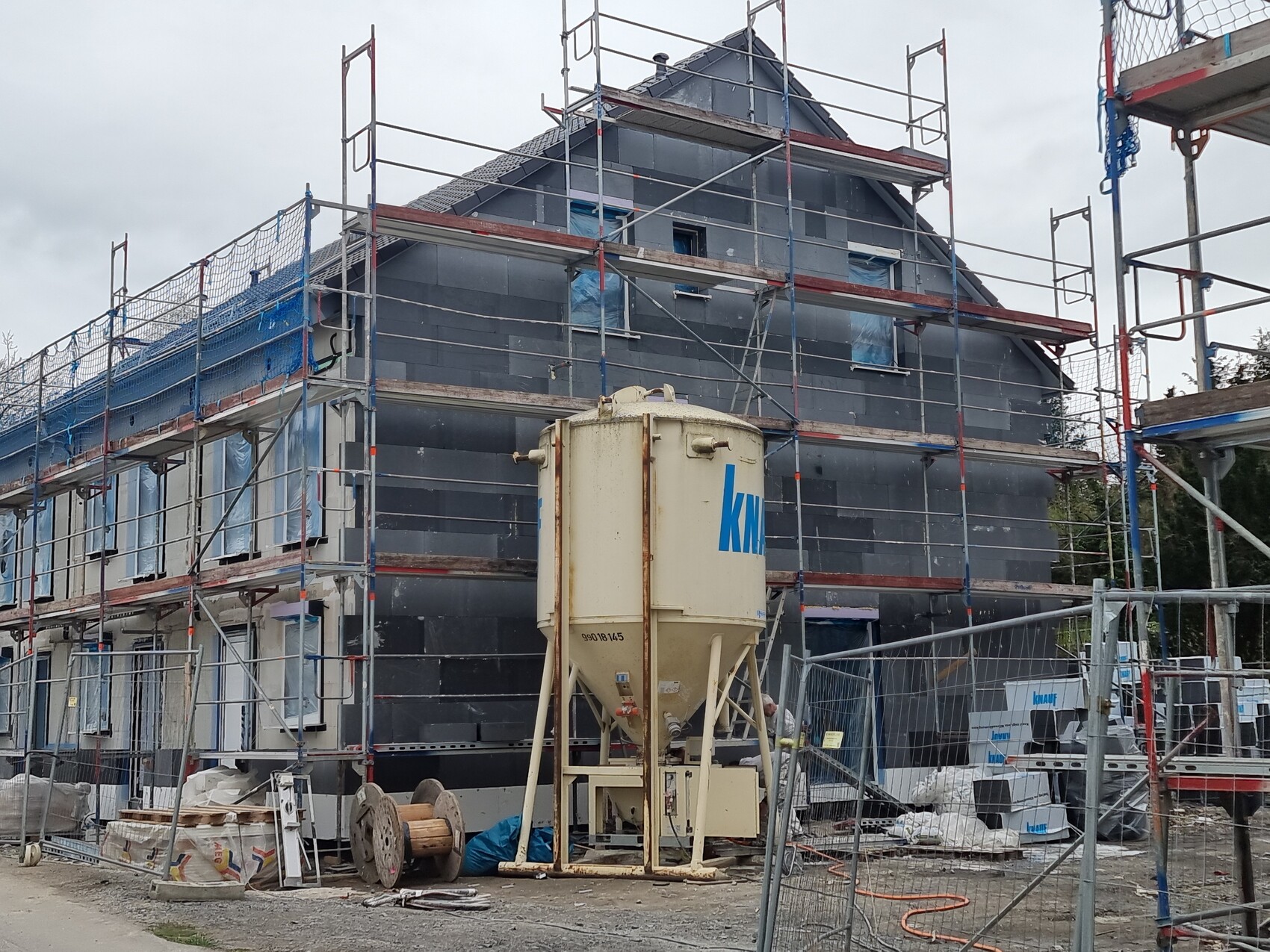 Zweigeschossiges Haus unter Gerüst mit blauen Abdeckungen; Betonmischer vorne.