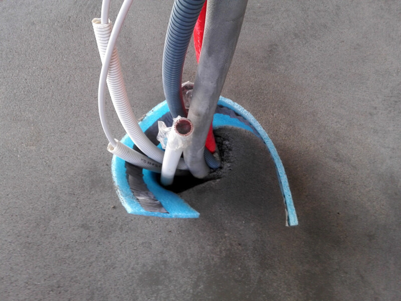 Offene Bodenbohrung im Rohbau mit Kabeln, Schläuchen und blauem Dämmmaterial.