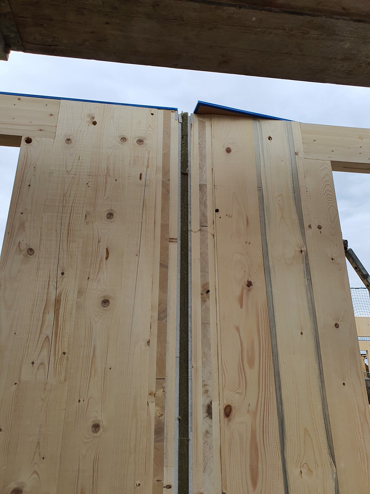 Rohbau: Vertikale Holzbretter bilden Wand, Spalte mit Dämmstoff, blauer Himmel sichtbar.