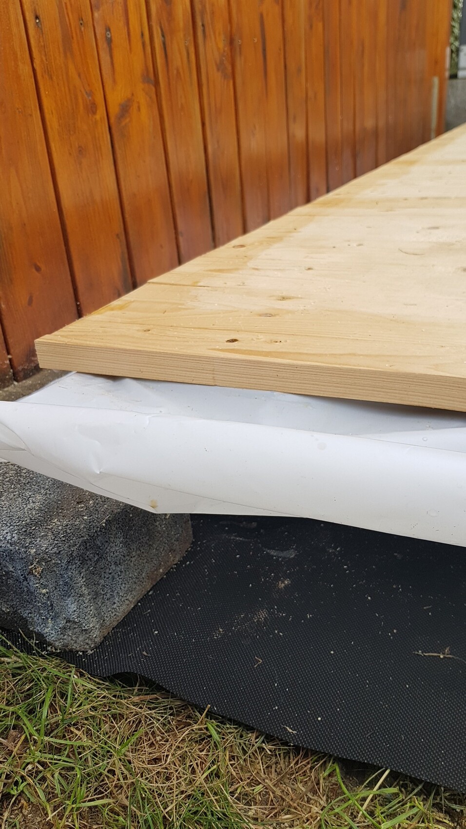 Eine Holzplatte liegt auf einer weißen Baufolie über dem Fundament neben einer vertikalen Holzwand.