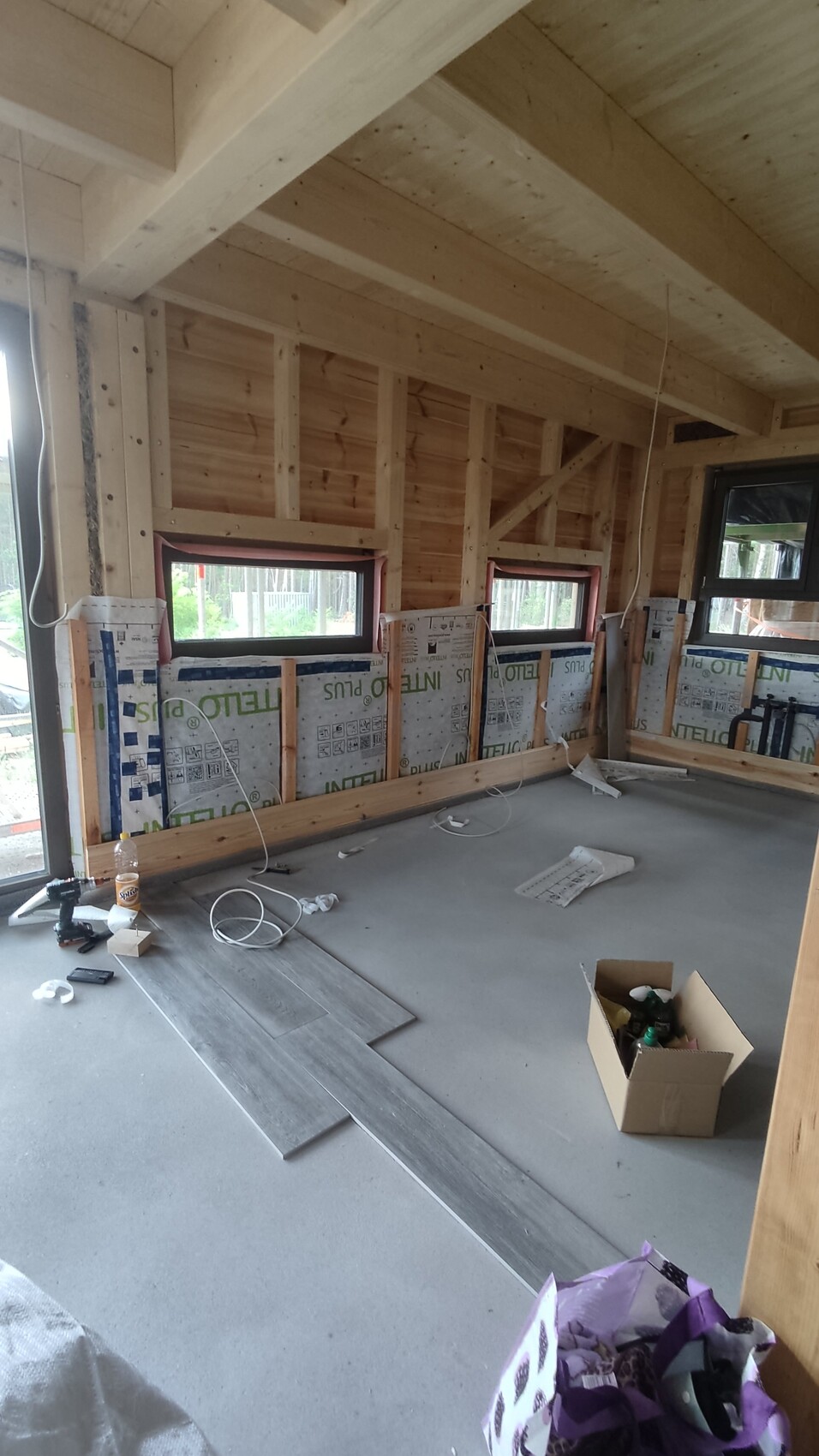 Innenraum im Rohbau: Holzständerwerk, Fenster, Dämmung und verstreutes Werkzeug auf Betonboden.