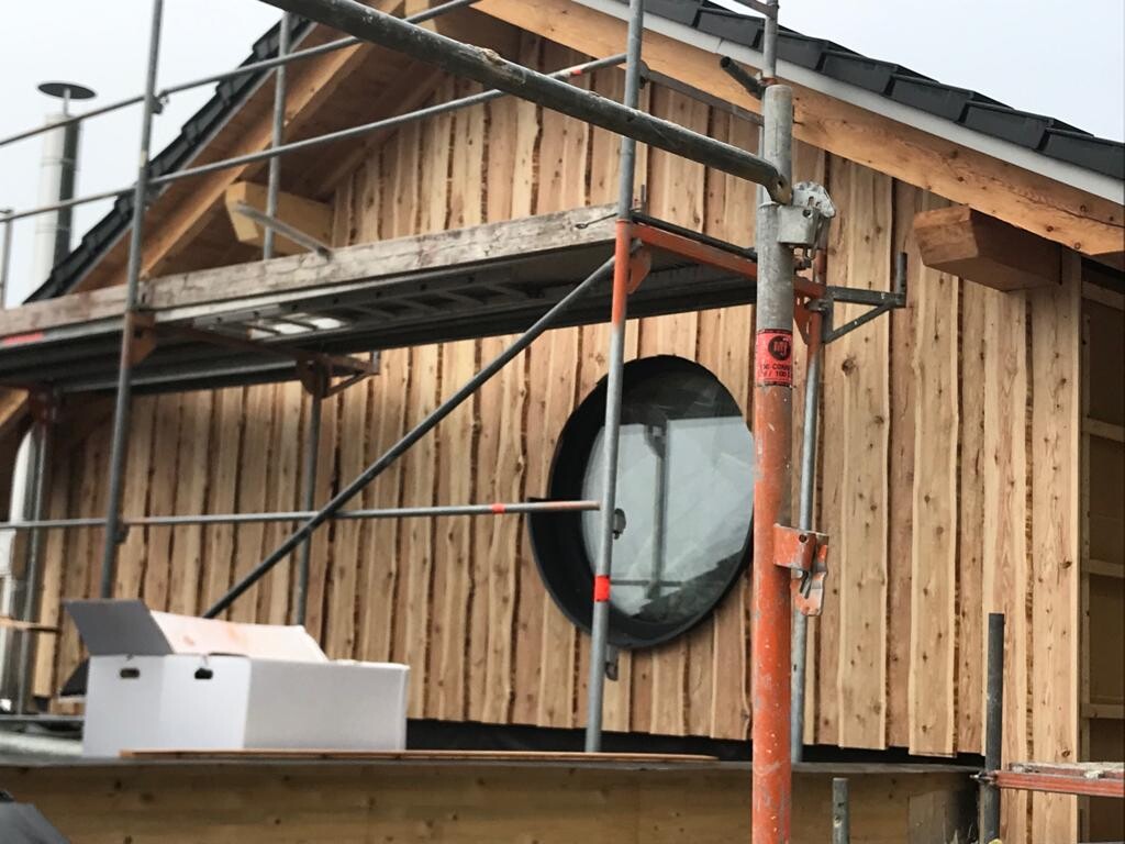 Baustelle: Holzfassade, orangefarbenes Gerüst und rundes Fenster am Neubau; Baukisten daneben.