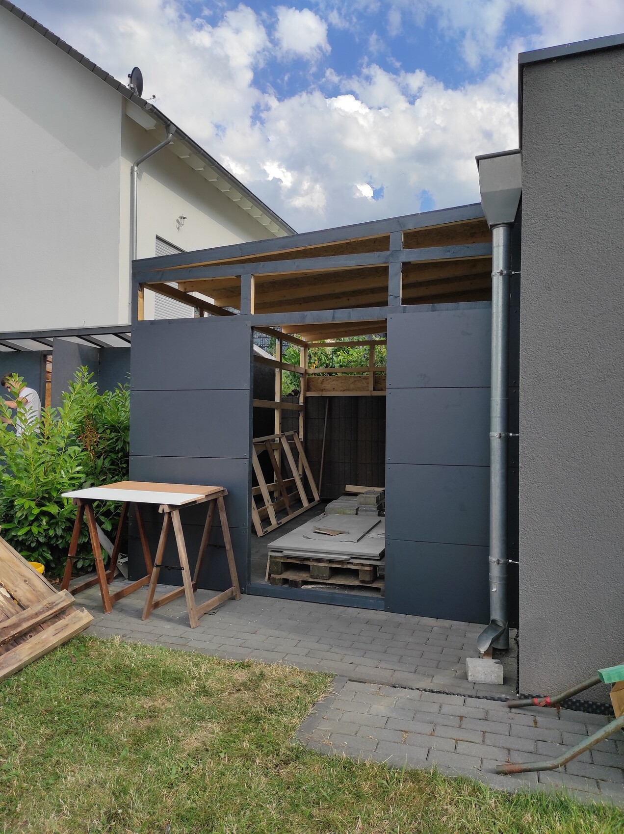 Bau im Garten: offenes Holz-Metall-Gerüst, dunkle Wandpaneele, Paletten.