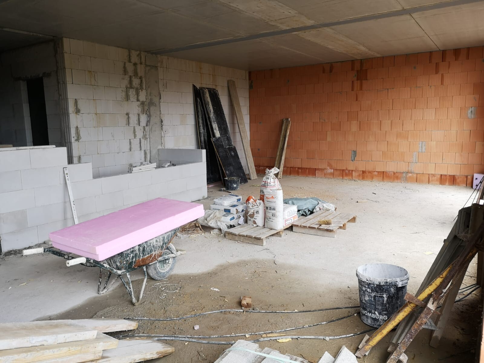 Baustelle: weiße Blöcke links, orange Ziegelwand rechts; Schubkarren mit pinkem Dämmblock.