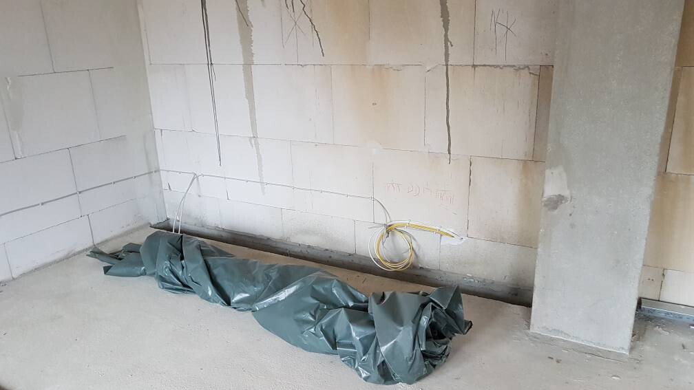 Unverputzter Bauraum: Ziegelbetonwand, Betonstütze, graue Folie auf dem Boden, gelbe Kabel.