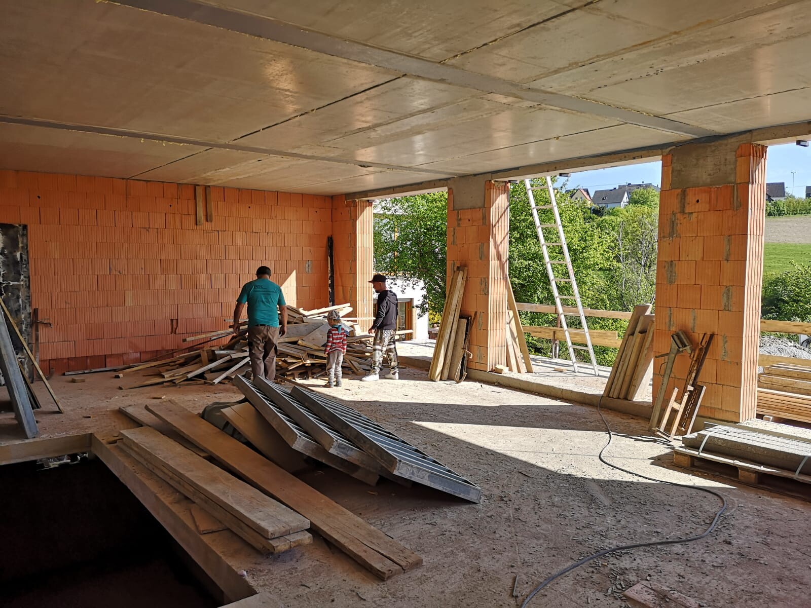 Rohbau eines Ziegelhauses drei Personen sortieren Holzreste, Leiter steht, Außen Grünland