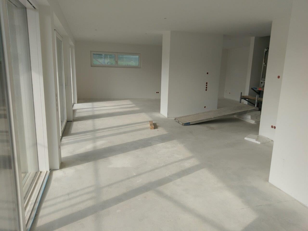 Großer heller Innenraum im Rohbau mit weißen Wänden, großen Fenstern und Baugeräten.