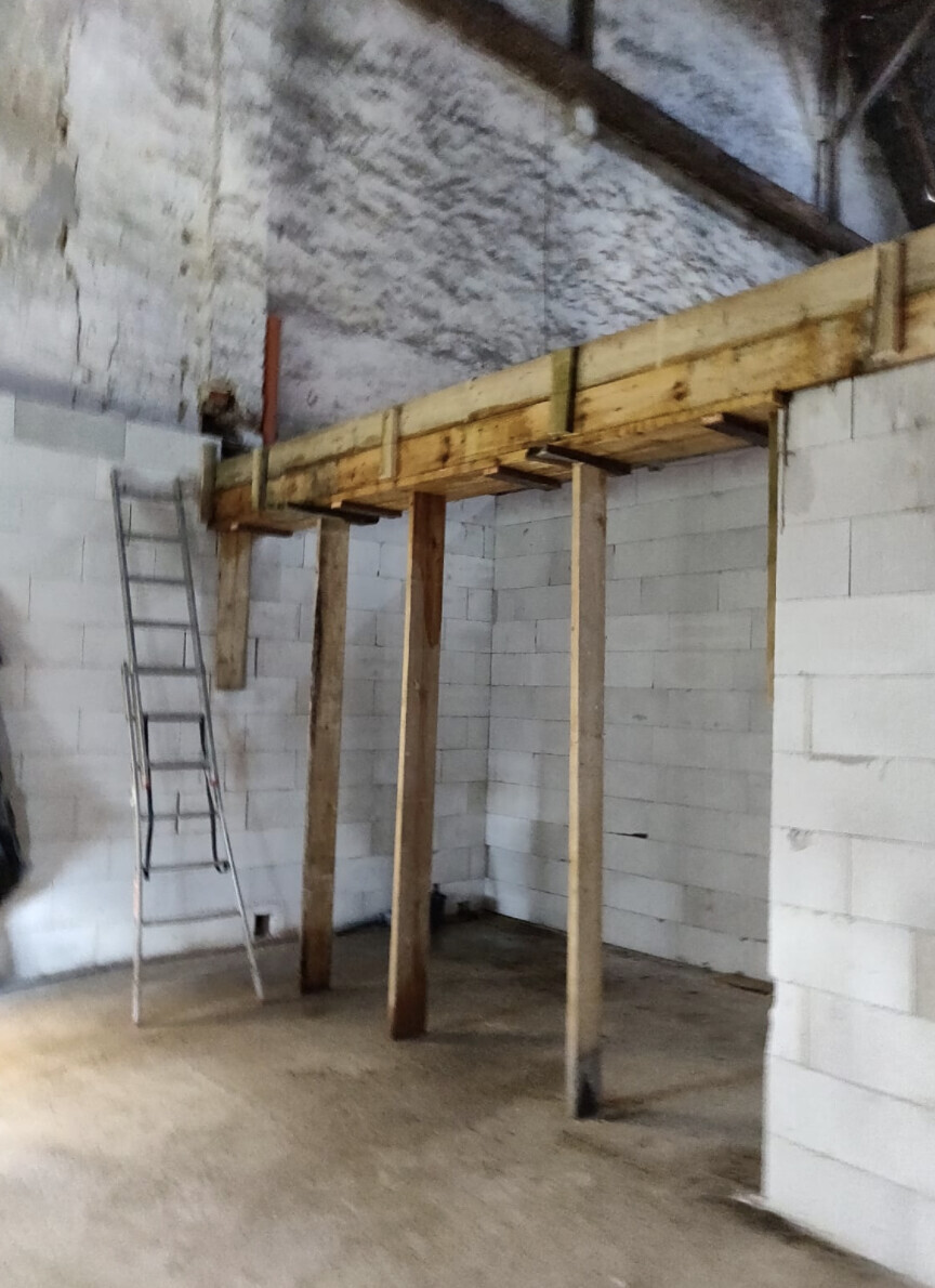 Innenraum einer Baustelle mit quer liegender Holzbalkenkonstruktion, drei Stützen und Leiter links.