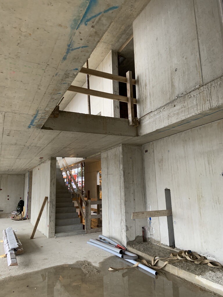 Rohbau: Betonwände, Treppe mit Holzgeländer, Bauwerkzeuge und Pfütze im Raum.