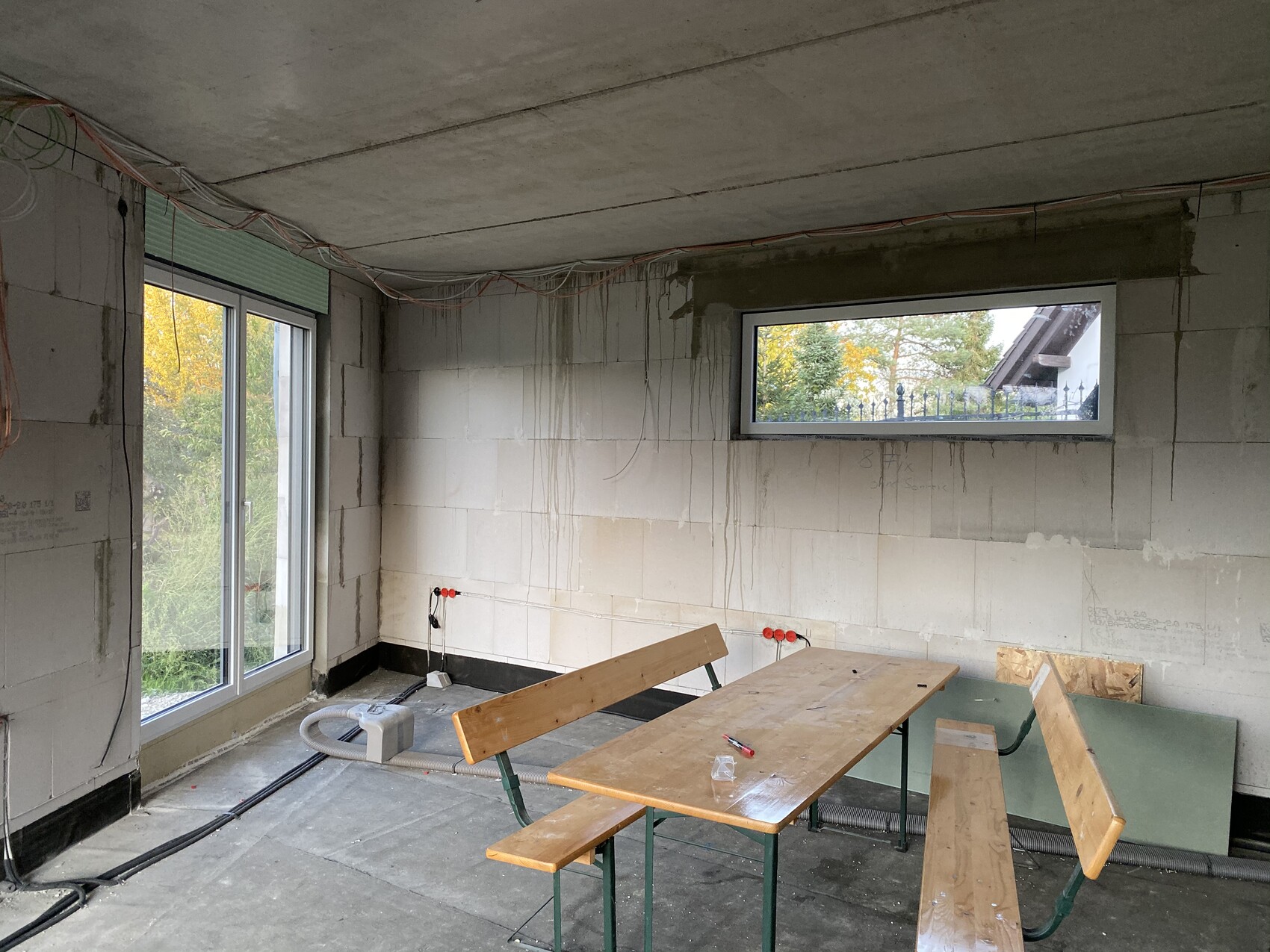 Unfertiger Bau-Raum mit Holztisch und Bänken, lose Kabel an Decke, Fenster mit Gartenblick.