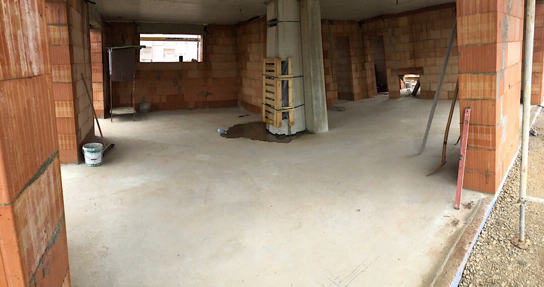Rohbau-Innenraum: Betonboden, Ziegelwände, Stützen und Baugeräte sichtbar.