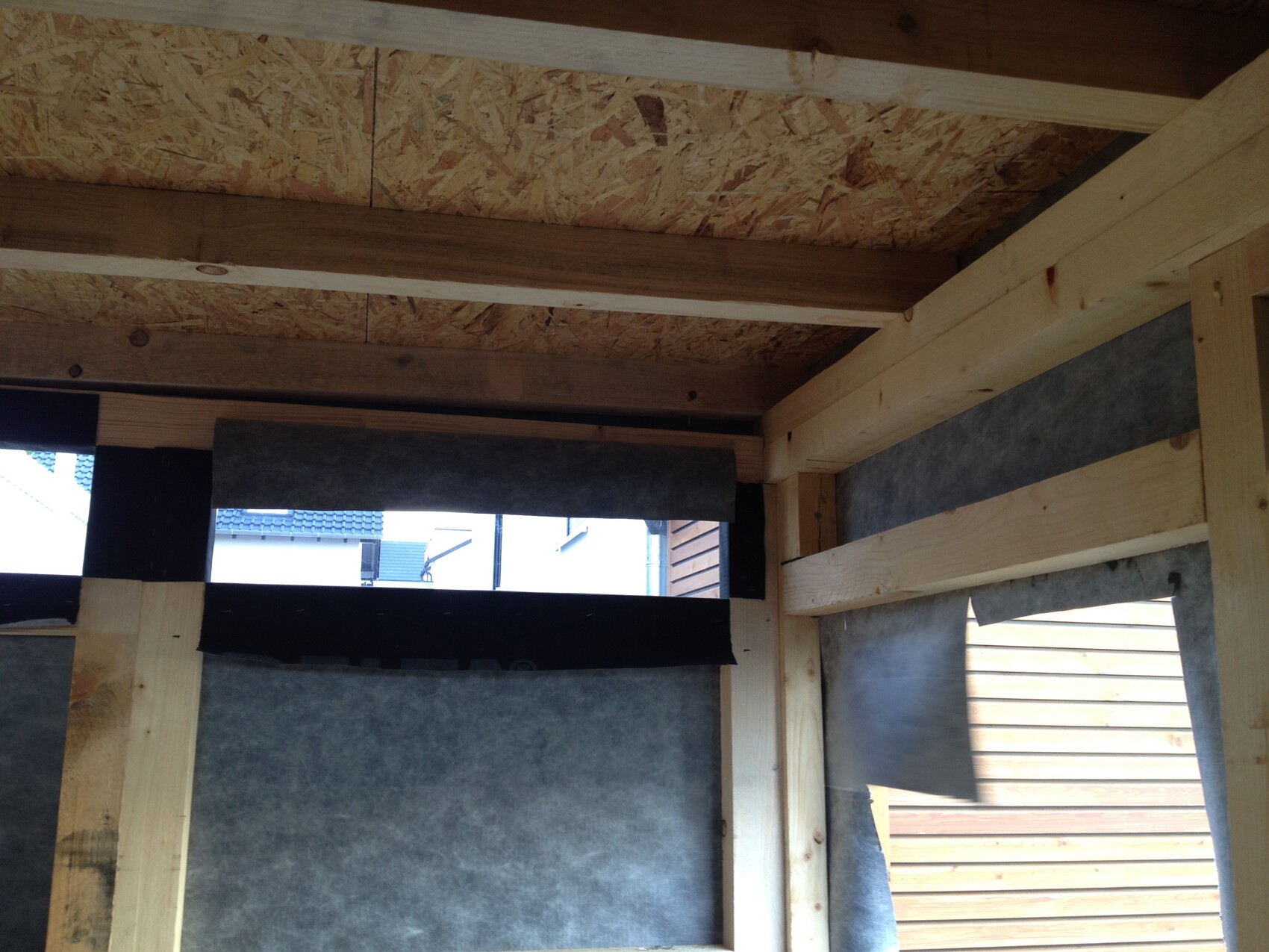 Innenraum eines Rohbaus: Holzbalken, OSB-Decke, Dämmplatten und Fensteröffnung sichtbar.