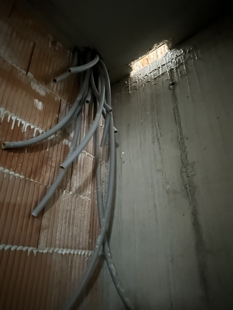 Baustellen-Ecke: graue Kabelschläuche hangen vor rohem Mauerwerk-links und Betonwand-rechts.