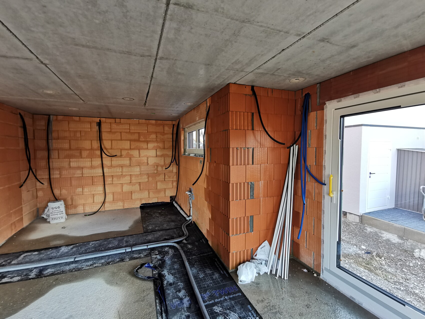 Unfertiger Innenraum mit Ziegelwänden, hängenden Kabeln, Rohren, Fenster und Glastür nach draußen