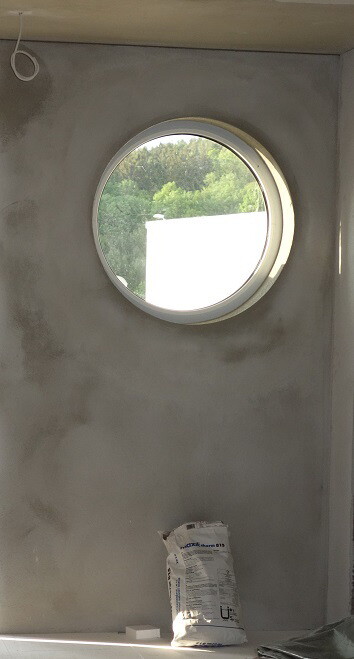 Rundes Fenster in rauer Wand einer Baustelle; Zementsack liegt auf dem Boden.