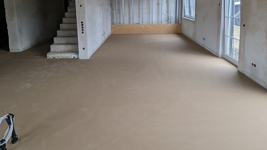 Offene Baustelle: roher brauner Boden, linke Treppe, Betonwände und große Fenster rechts.