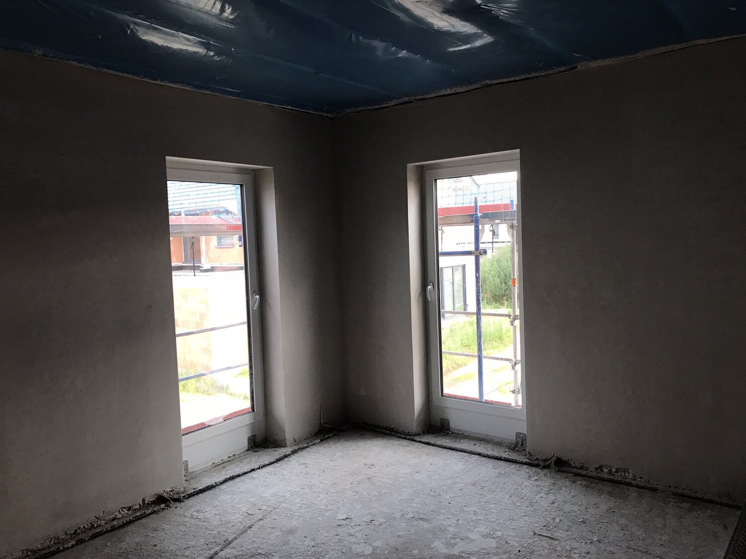 Innenraum im Rohbau mit zwei Fenstern, unverputzte Wände und Baugerüst sichtbar