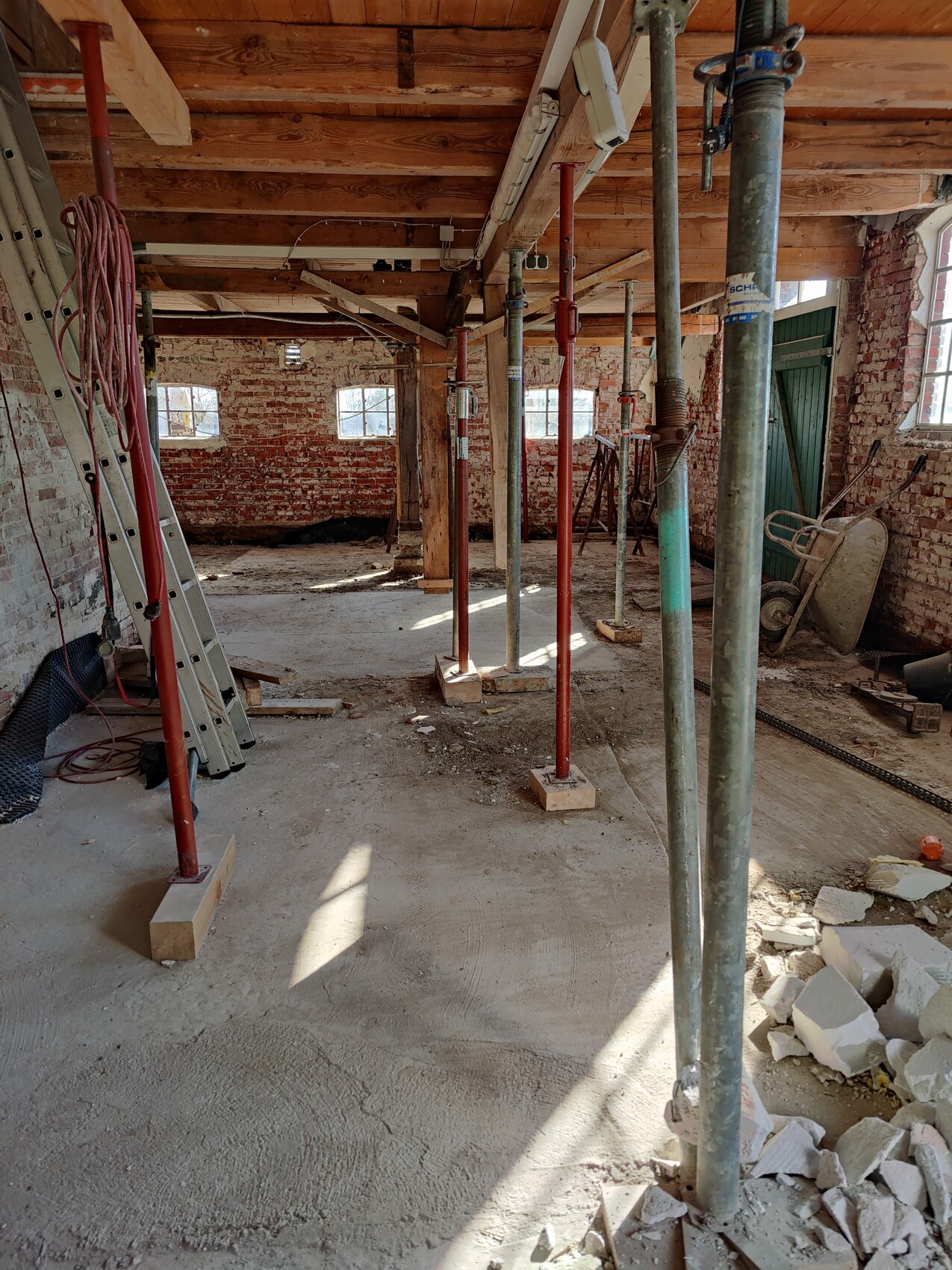 Baustellenraum mit roten Stützstangen, Holzbalkendecke, Bruchsteinwänden, Schutt und Werkzeug.
