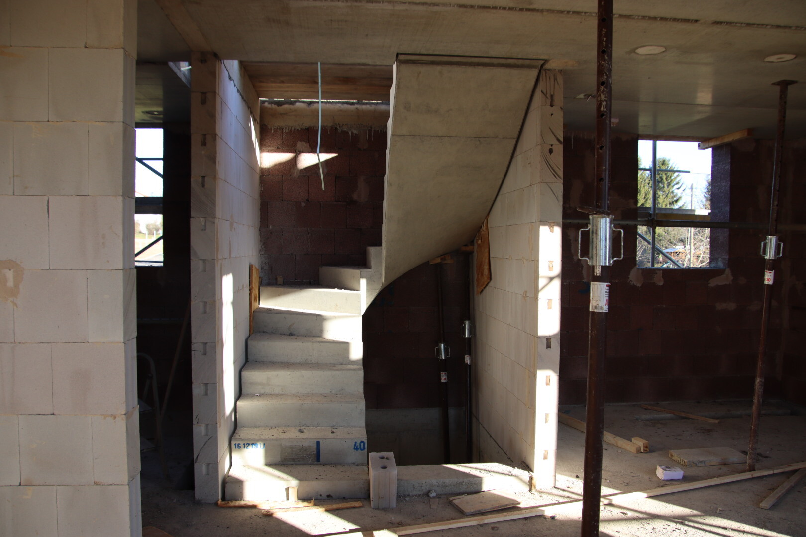 Innenraum eines Rohbaus mit abgerundeter Betontreppe, Blockwänden, Stützen und Baugerüsten.